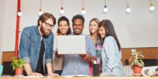 Team gathered around a laptop, smiling and collaborating in a brightly lit office.