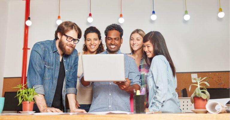 Team gathered around a laptop, smiling and collaborating in a brightly lit office.