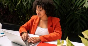 Woman in an orange blazer working on a laptop at an outdoor table.