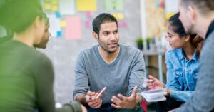 Small group in a meeting, with one man speaking and gesturing while others listen and take notes.