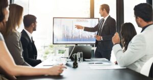 Business presentation with a man pointing to a bar graph on a screen in a meeting room.