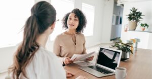 Two businesswomen having a discussion at a desk with a laptop and papers.