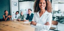 Smiling businesswoman standing confidently in front of a meeting table with colleagues.