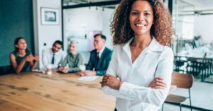 Smiling businesswoman standing confidently in front of a meeting table with colleagues.
