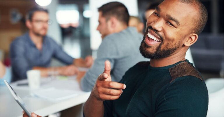 Smiling man pointing while holding a tablet in an office meeting.