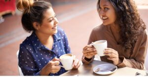 Two women smiling and chatting over coffee at a cafe table.