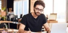 Man in glasses writing at a desk with a laptop.