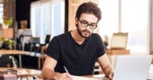 Man in glasses writing at a desk with a laptop.