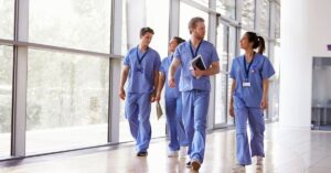Group of healthcare professionals in scrubs walking through a bright hospital corridor, engaged in conversation and holding clipboards and tablets.