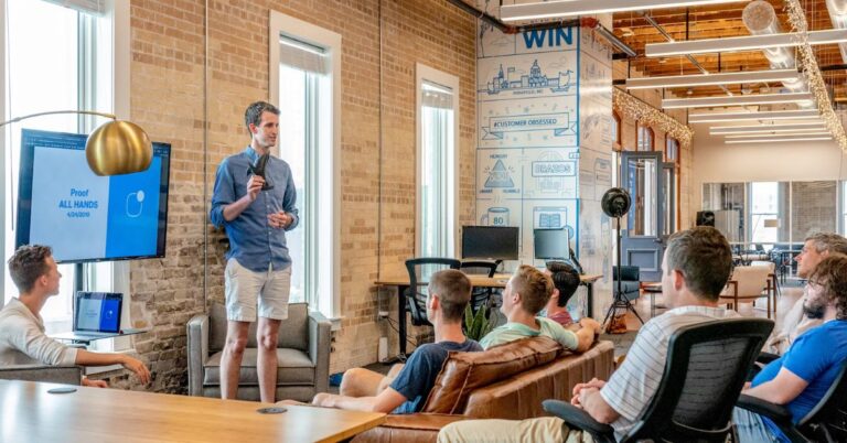 A group of people in a modern office listen to a man giving a presentation, with a screen showing “Proof ALL HANDS 4/24/2019” in the background.