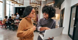 Two colleagues smiling and discussing work on a laptop in a modern office.