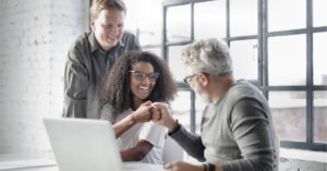 Three coworkers fist-bumping and smiling around a laptop in a bright office.