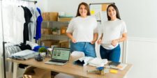 Two women in a clothing workspace, smiling with folded T-shirts and a laptop displaying an online store.