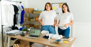 Two women in a clothing workspace, smiling with folded T-shirts and a laptop displaying an online store.