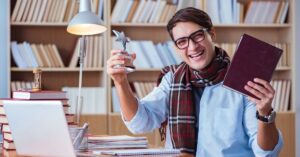 Smiling man wearing glasses and a scarf, holding a book in one hand and a small star-shaped trophy in the other, sitting at a desk with books and a laptop.