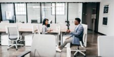 Host filming a guest for a workplace vlog in a glass-partitioned office.
