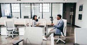 Host filming a guest for a workplace vlog in a glass-partitioned office.