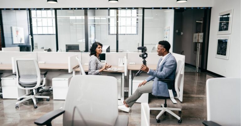 Host filming a guest for a workplace vlog in a glass-partitioned office.
