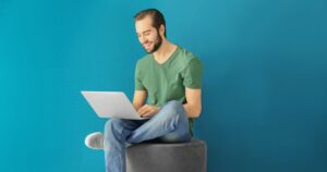 Smiling man in a green shirt sitting cross-legged on a stool, working on a laptop against a bright teal background.