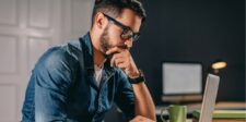 Man wearing glasses focused on a laptop, hand on chin, deep in thought.