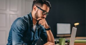 Man wearing glasses focused on a laptop, hand on chin, deep in thought.