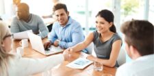 Smiling businesswoman shaking hands across a meeting table while colleagues look on and a laptop sits open.