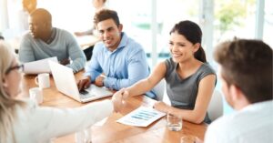 Smiling businesswoman shaking hands across a meeting table while colleagues look on and a laptop sits open.