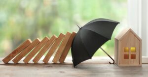 A row of wooden blocks toppling toward a small house model is stopped by a black umbrella, symbolizing insurance protection and risk management.