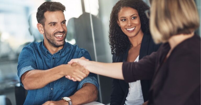 Two colleagues smiling while one shakes hands with another person in an office setting.