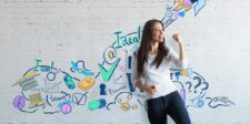 Smiling woman standing in front of a white brick wall decorated with colorful hand-drawn graphics of books, charts, a rocket, and other business symbols, representing ideas, success, and growth.