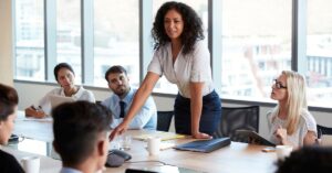 Businesswoman leading a team meeting, speaking to colleagues seated around a conference table in a modern office.