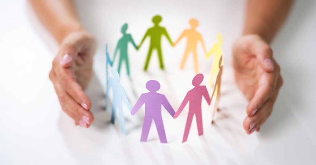 Hands surrounding a circle of colorful paper cut-out figures holding hands, symbolizing diversity and inclusion.