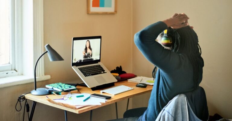 A person working from home sits at a desk with a laptop on a video call, hands on their head, wearing colorful headphones, with notebooks and papers scattered around.