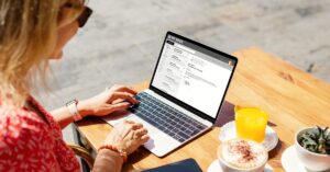 Woman sitting at an outdoor table using a laptop with her email inbox open, next to orange juice, coffee, and a small plant.