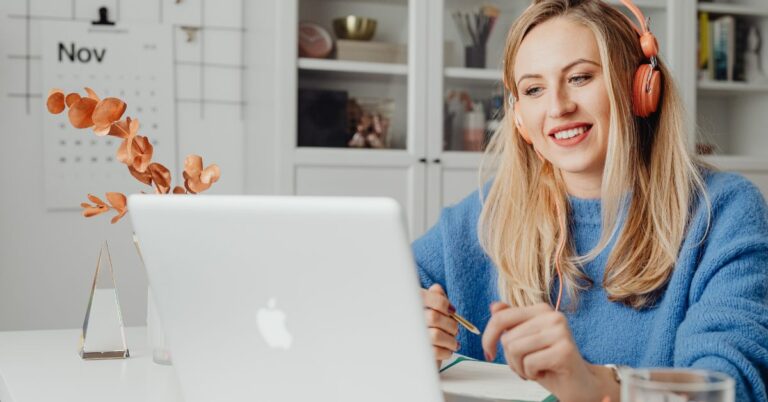 Young woman with headphones working on a laptop at home, smiling during remote work with a notebook in front of her.