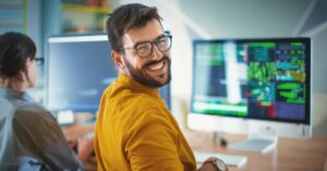 Smiling male developer in glasses and yellow shirt sitting at desk with computer monitors displaying code, while colleague works beside him.