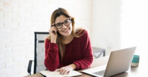Smiling woman wearing glasses and a red sweater sitting at a desk with a laptop and notebook, looking confident and ready to write.