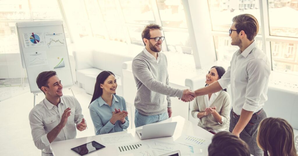 Team of professionals in a bright modern office celebrating a successful business deal as two colleagues shake hands while others applaud around a conference table.