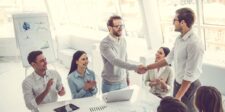 Team of professionals in a bright modern office celebrating a successful business deal as two colleagues shake hands while others applaud around a conference table.