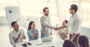 Team of professionals in a bright modern office celebrating a successful business deal as two colleagues shake hands while others applaud around a conference table.