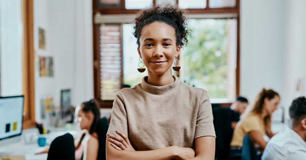 Confident young professional standing in a modern office with design team working in the background.