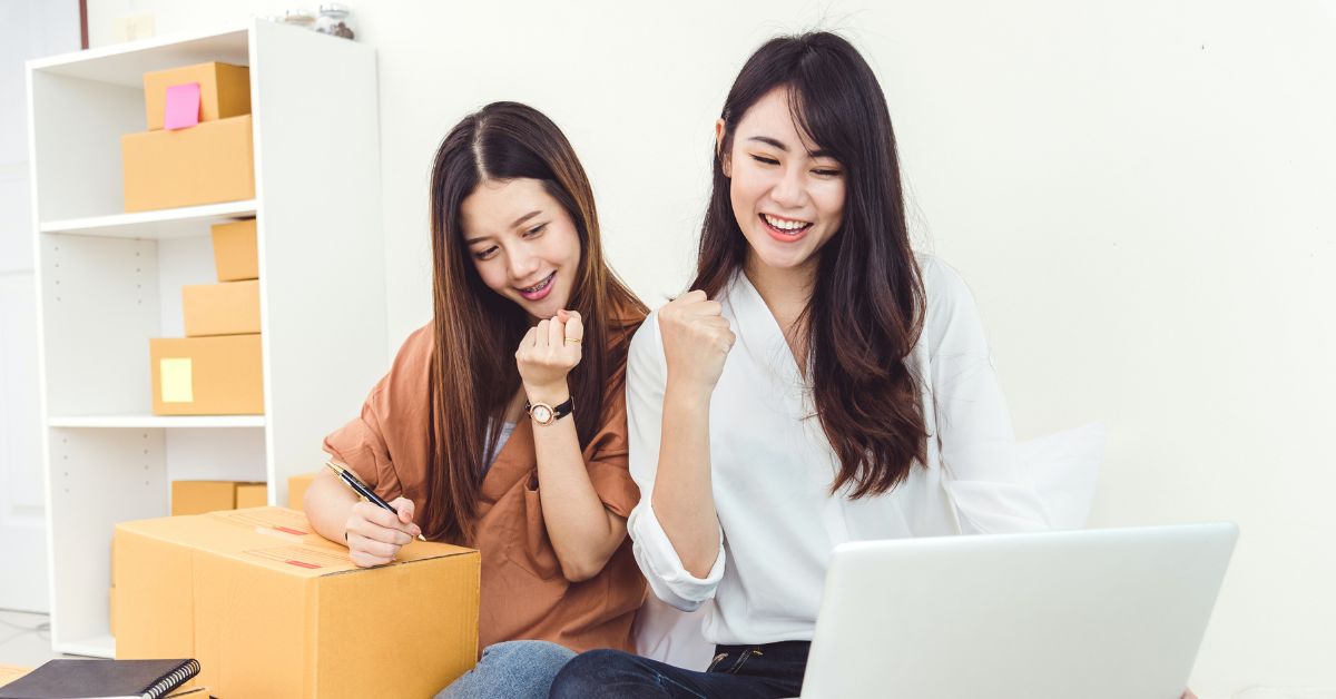 Two happy young entrepreneurs celebrating success while working on an online business with boxes and a laptop nearby.