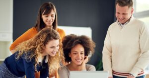 Four colleagues gathered around a laptop, smiling and looking at the screen together in an office.