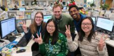 Smiling office team posing at their desks in a decorated open-plan workspace, holding up peace signs in front of computer screens.