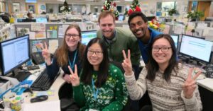 Smiling office team posing at their desks in a decorated open-plan workspace, holding up peace signs in front of computer screens.
