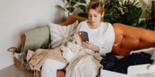 Woman sitting on a tan couch surrounded by clothes, focused on her smartphone—used to illustrate how busy customers browse apps and websites in everyday life and expect them to work smoothly.