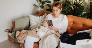 Woman sitting on a tan couch surrounded by clothes, focused on her smartphone—used to illustrate how busy customers browse apps and websites in everyday life and expect them to work smoothly.