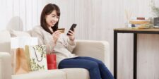 Smiling woman sitting on a couch with shopping bags, holding a credit card and smartphone while shopping online, representing a positive customer experience.