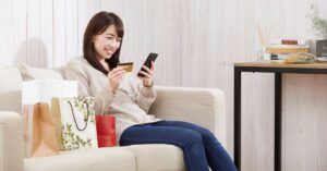 Smiling woman sitting on a couch with shopping bags, holding a credit card and smartphone while shopping online, representing a positive customer experience.