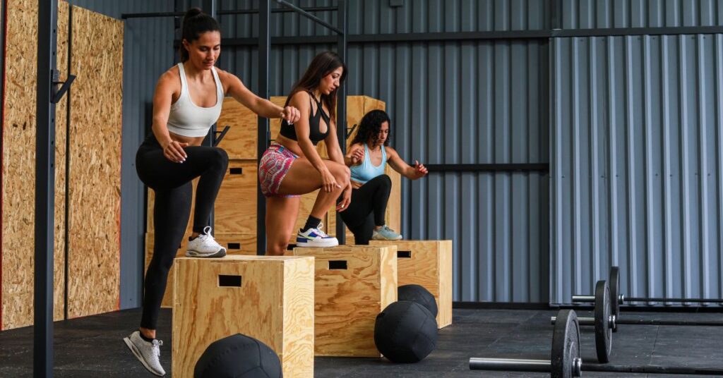 Three women performing box step-ups during a CrossFit workout inside a modern gym. Wooden plyo boxes, barbells, and medicine balls are visible on the floor, with metal panel walls in the background.